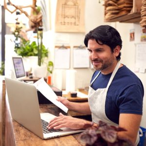Male Sales Assistant Working On Laptop Behind Sales Desk Of Florists Store